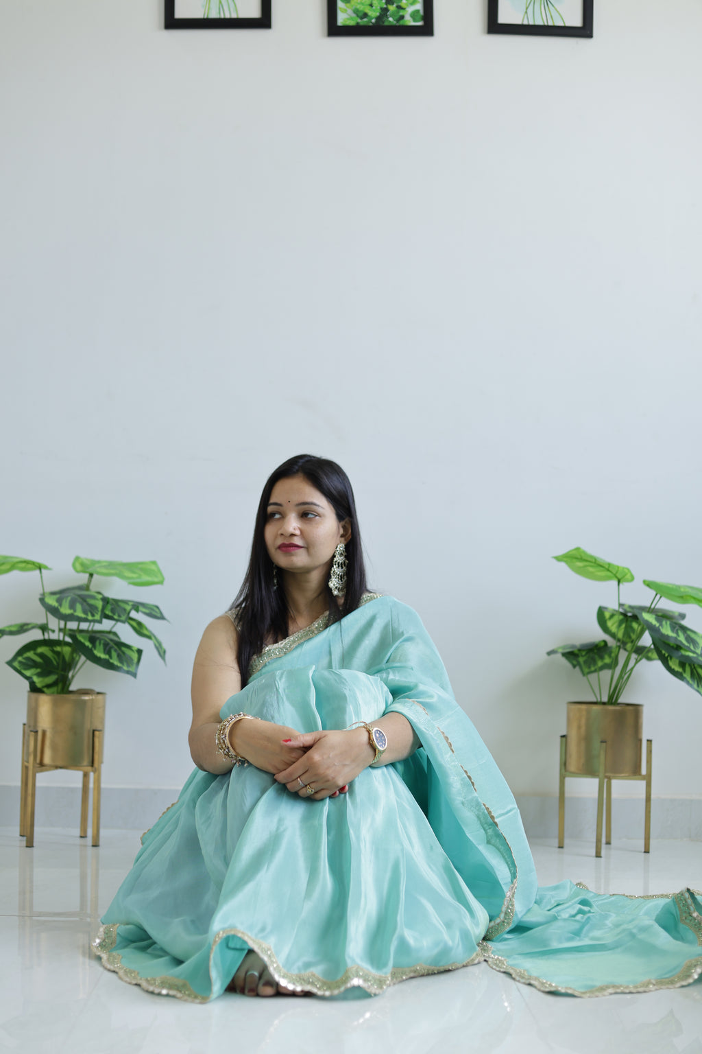 Woman in a light blue saree sitting on a white floor with plants in the background