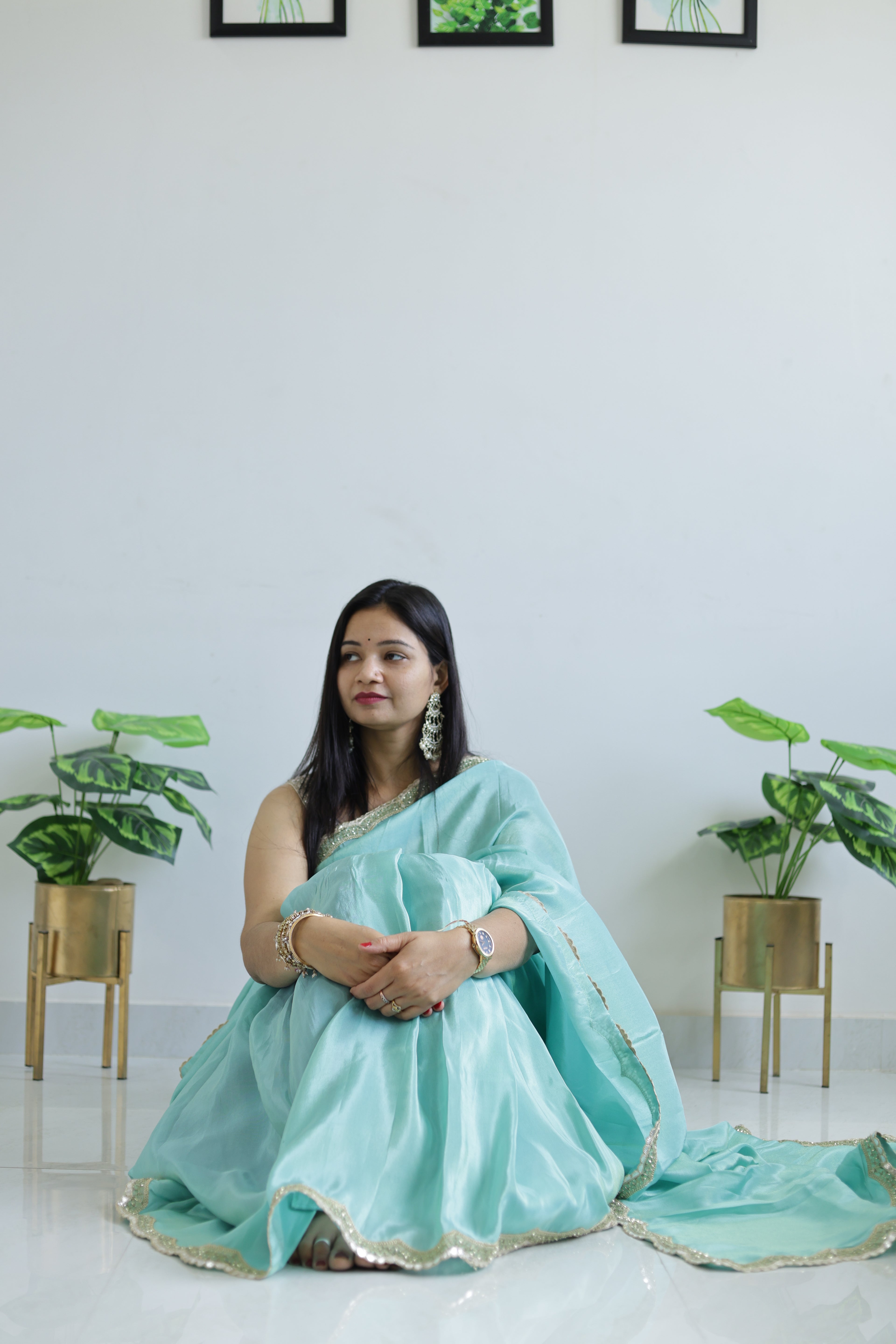 Woman in a light blue saree sitting on a white floor with plants in the background